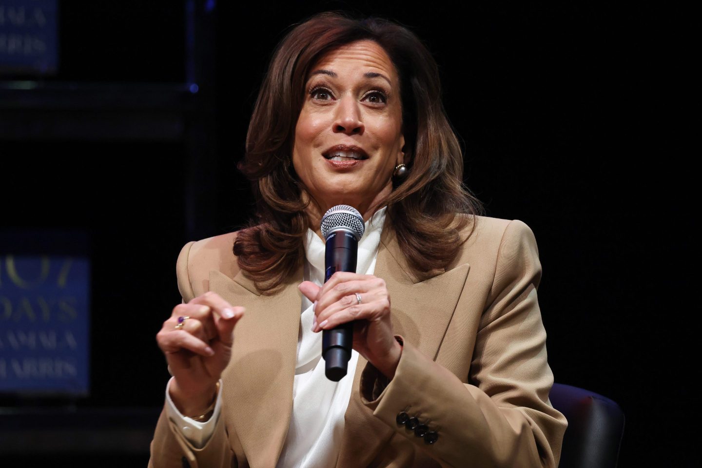 A seated woman in a tan suit jacket speaks into a handheld microphone on stage. A book cover is enlarged behind her